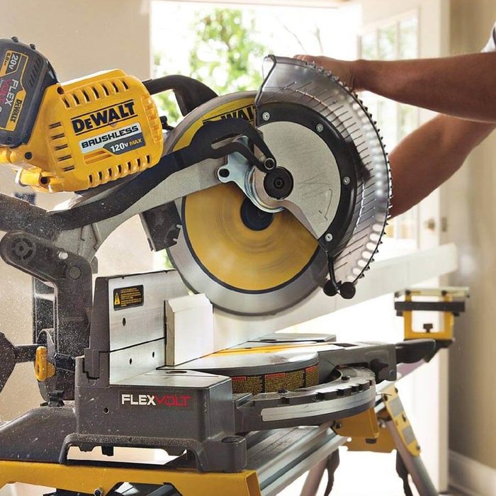 A man operates a miter saw, cutting a piece of wood, in a well-lit room with windows and light-colored walls. Dust particles fill the air.