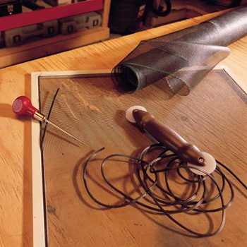 A screen frame rests on a wooden table, with a roll of mesh nearby, a pointed tool, and a brown handle with attached black cords.