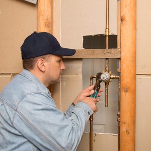 A plumber is using a wrench to adjust pipe fittings inside a wall cavity, surrounded by wooden framing and drywall.