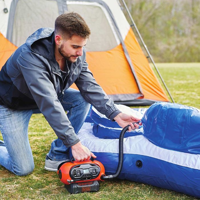 A man uses an air pump to inflate a blue sleeping pad while kneeling on grass near an orange and gray tent setup in the background.