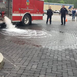 A fire truck discharges water onto a paved area, creating ripples. Two uniformed personnel observe, while others are visible in the background.