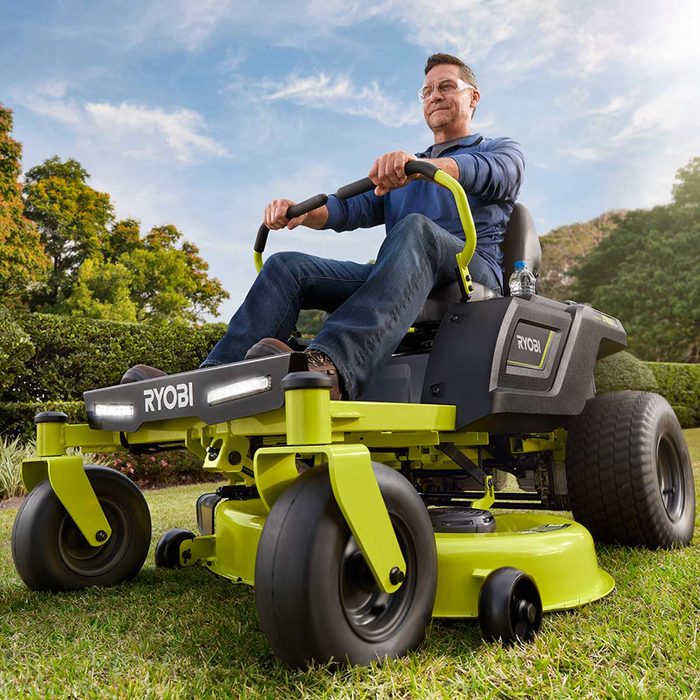 A man operates a riding lawn mower on grass, surrounded by shrubs and trees under a clear blue sky, showcasing a well-maintained yard.