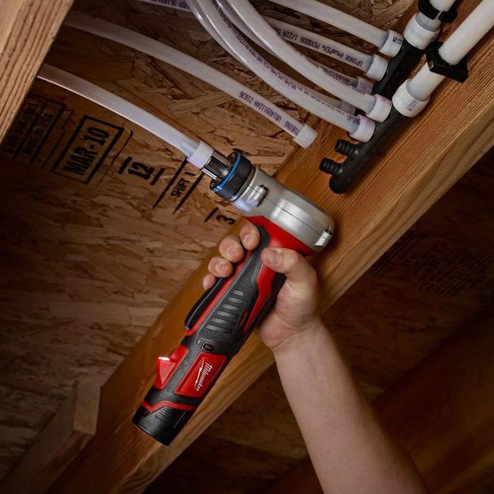 A hand holds a red and silver tool, attaching it to white tubes mounted under a wooden structure in a dimly lit environment.