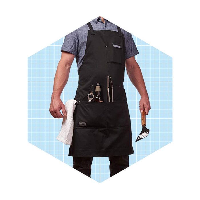 A person wearing a black apron holds a spatula and tongs while displaying various tools in the apron pockets against a light blue grid background.