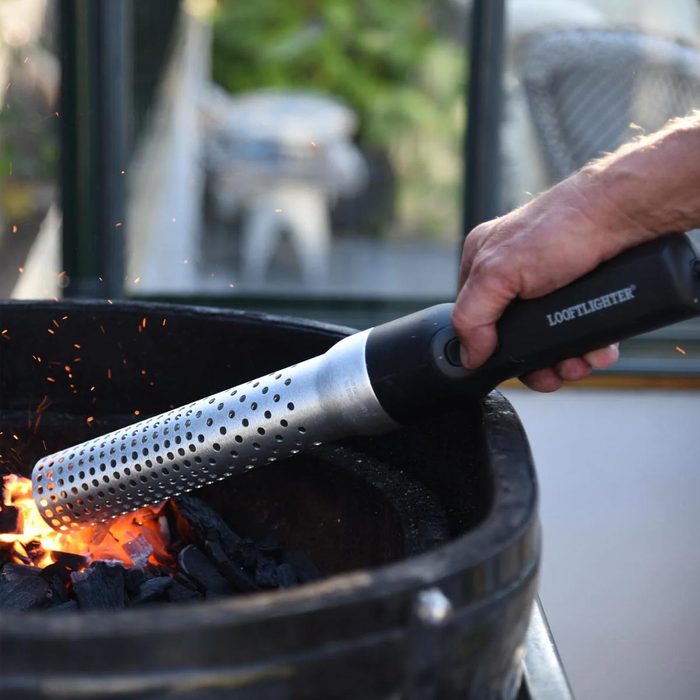 A metal firestarter is igniting charcoal in a grill, producing sparks in a backyard setting, with a blurred background suggesting outdoor furniture.