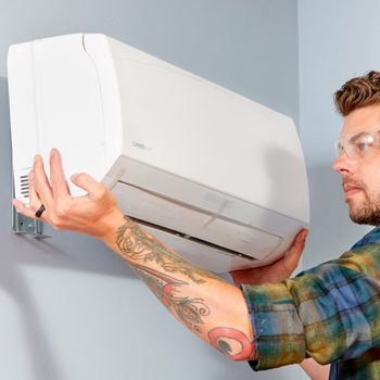 A person installs a wall-mounted air conditioner, focused and wearing safety glasses, in a blue-painted room. The individual's arm showcases a detailed tattoo.