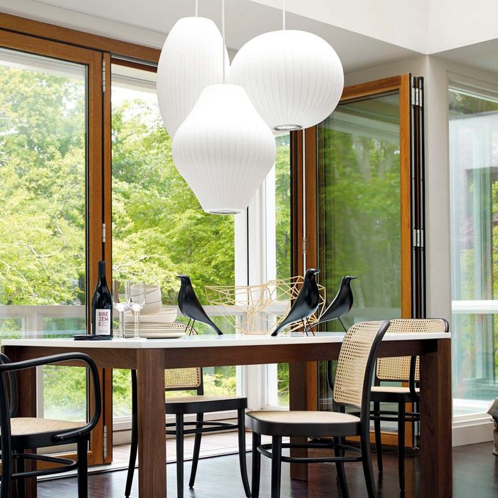 Three white pendant lights hang above a wooden dining table, which features black bird figurines and a wine bottle, surrounded by large windows showcasing greenery outside.