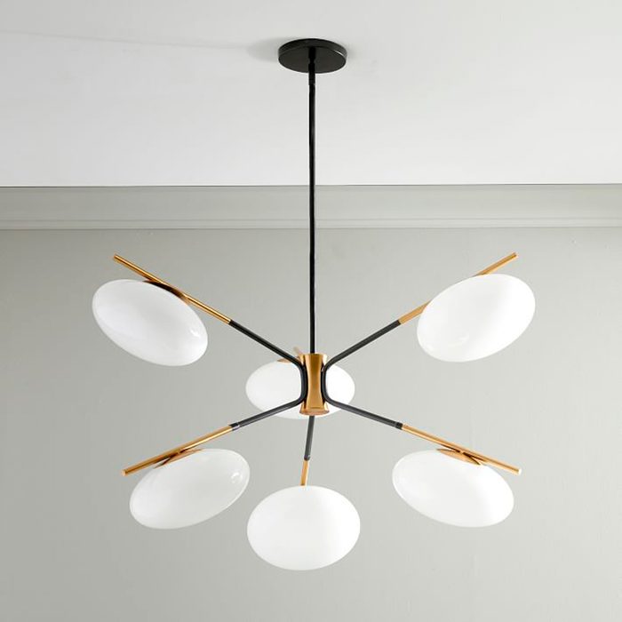 A modern chandelier with six white glass shades extends from a central black and gold fixture, hanging from the ceiling in a minimalist room.