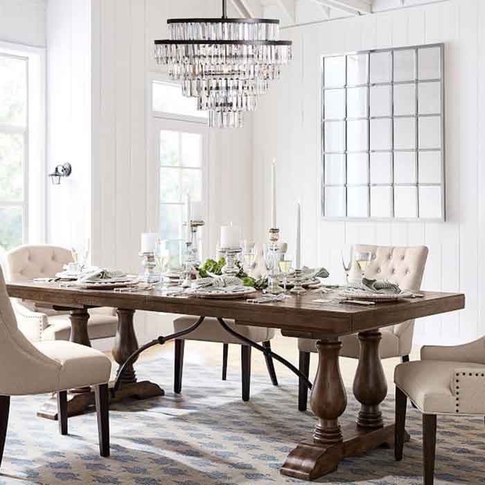 A wooden dining table is set with elegant dinnerware and candles, surrounded by upholstered chairs, in a bright room with a chandelier and mirror.