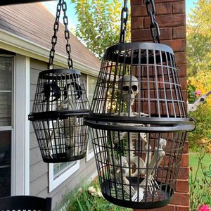 Two black wire cages hang from a porch, each containing a skeleton. The background features a house and greenery with flowers.