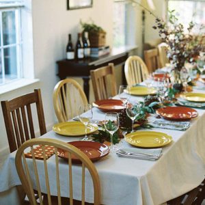 A beautifully set dining table displays colorful plates and glasses, surrounded by wooden chairs, with a warm, inviting atmosphere enhanced by natural light.