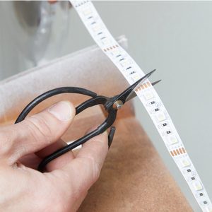 A hand uses scissors to cut a flexible LED strip, positioned above a brown surface, indicating a task related to lighting installation or adjustment.