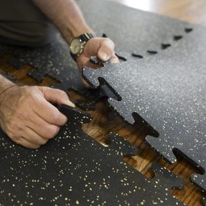 A person is fitting interlocking black rubber puzzle pieces together on a wooden floor. The pieces have a speckled texture.