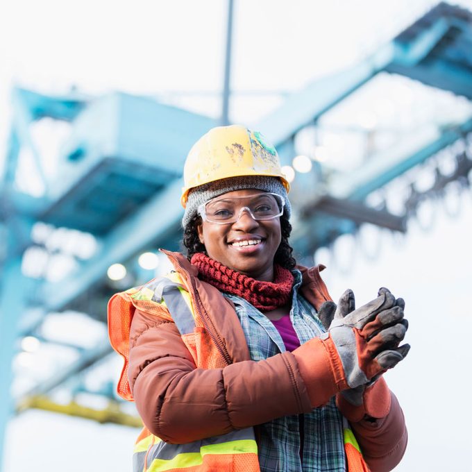 A woman in a hard hat and safety gear smiles while clapping her hands, standing near a large industrial crane against a bright sky.