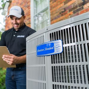 A technician examines a clipboard while standing near an American Standard air conditioning unit, surrounded by greenery and a brick wall.