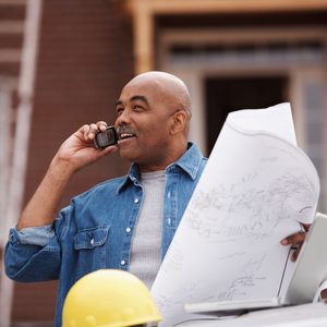 A man in a denim shirt holds blueprints while talking on a phone, standing near construction equipment outdoors with a building in the background.