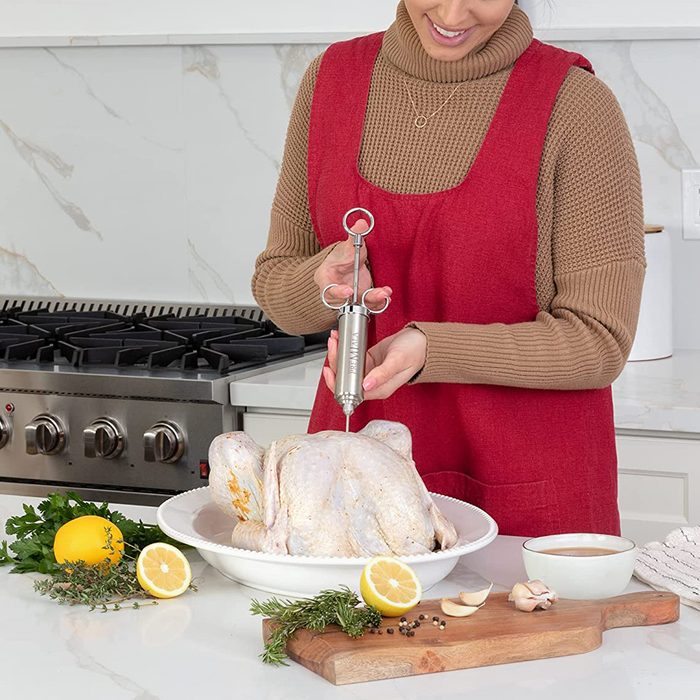 A person injects marinade into a raw turkey using a stainless steel baster, surrounded by herbs, lemons, and cooking utensils in a kitchen setting.