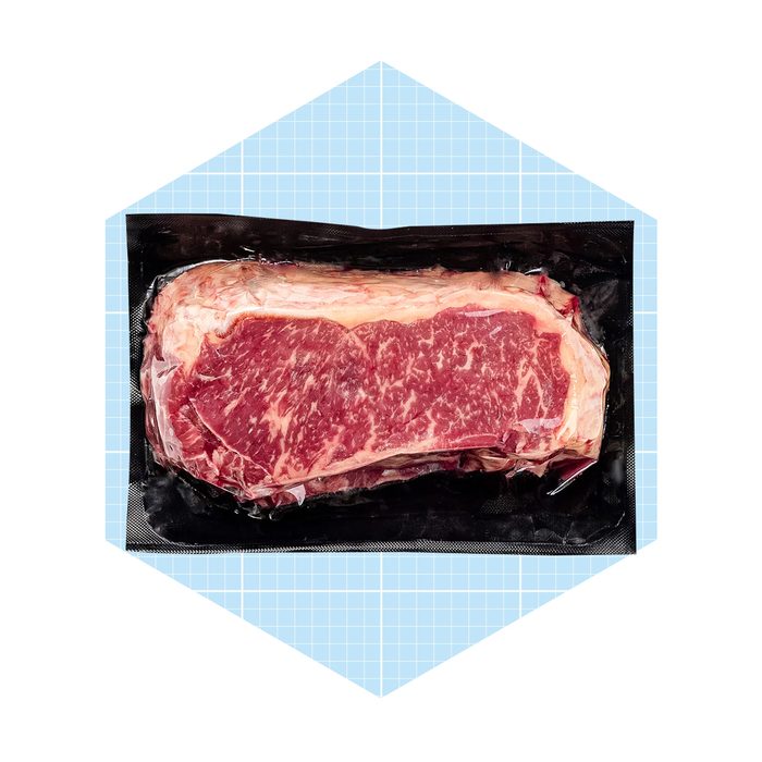 A raw steak is vacuum-sealed in a black plastic package, resting on a light blue grid-patterned background.