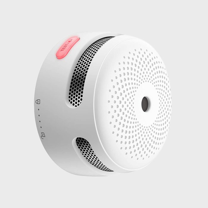 A round, white smoke detector features vents and a test button on top, mounted against a light gray background, indicating its safety monitoring function.