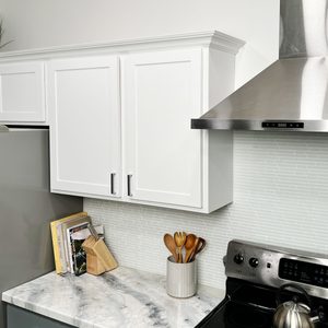 A marble countertop hosts a cup of wooden utensils, cookbooks, and a stainless steel stove, with white cabinets and a sleek range hood above.