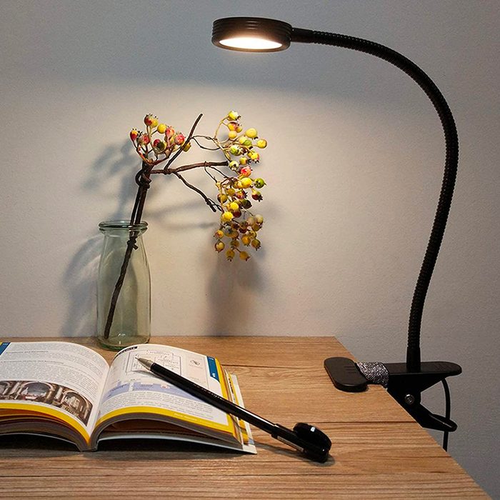 A desk lamp illuminates an open book on a wooden table, beside a glass vase holding a flower branch and a pen nearby.