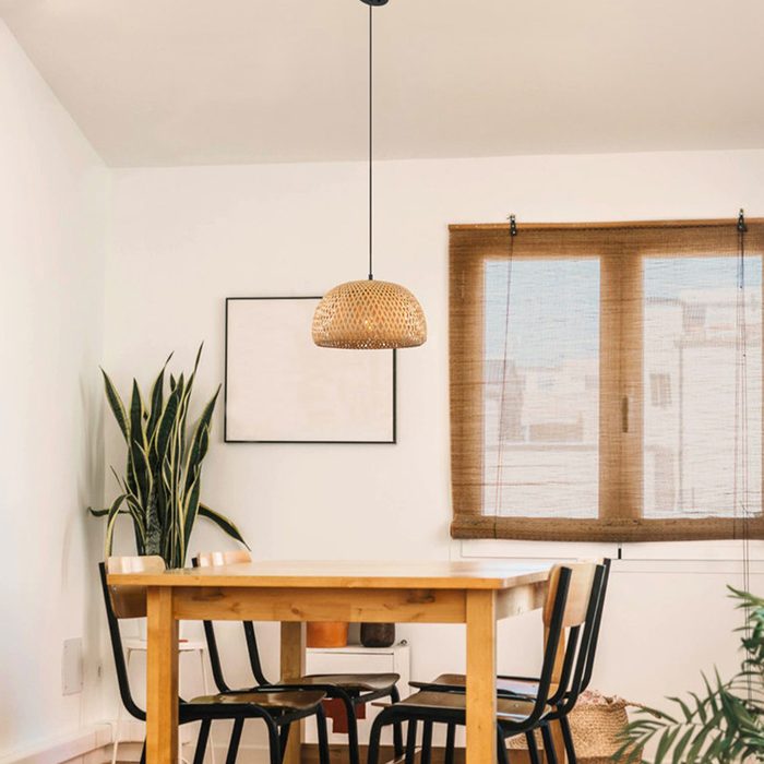 A wooden table with black chairs sits in a bright room, illuminated by a woven pendant lamp, with a plant and window nearby.