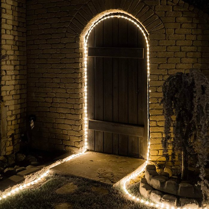 A wooden door, illuminated by string lights, stands in an arched brick entryway, surrounded by a stone pathway and a small potted plant.