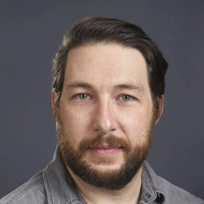 A man with dark hair and a beard faces the camera, wearing a gray shirt against a neutral background. His expression appears calm and focused.