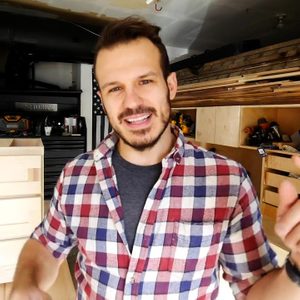 A man in a checkered shirt gestures while speaking in a workshop filled with wood, tools, and storage, emphasizing a wooden drawer project.