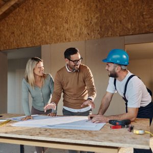 Three individuals examine construction plans on a table, smiling and engaging in discussion within a room with wooden beams and unfinished walls.