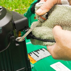 A person checks the oil level in a lawn mower engine using a dipstick while holding a cloth in a grassy outdoor setting.