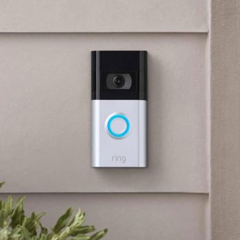 A doorbell camera mounted on a neutral-colored wall captures visitors, featuring a circular blue button labeled "ring" and surrounded by greenery.