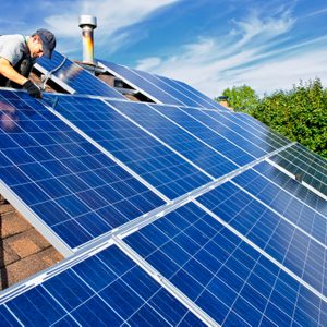 A worker is installing solar panels on a sloped roof under a clear blue sky, surrounded by green trees in the background.