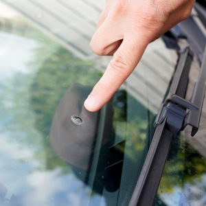 A hand points at a small crack on a car windshield, highlighting potential damage in a reflective, outdoor setting with greenery visible.