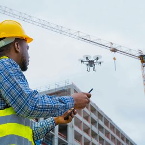 A construction worker operates a drone with a smartphone near a building site, featuring cranes and incomplete structures against a cloudy sky.