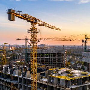 Cranes tower over partially constructed buildings, lifting materials against a backdrop of an urban skyline at sunset, with scattered trees and roads below.