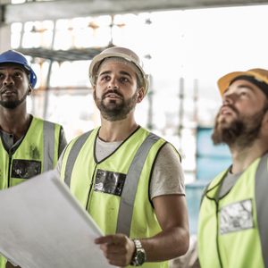 Three construction workers in safety vests examine blueprints while looking upward at a building site under construction, surrounded by scaffolding and natural light.