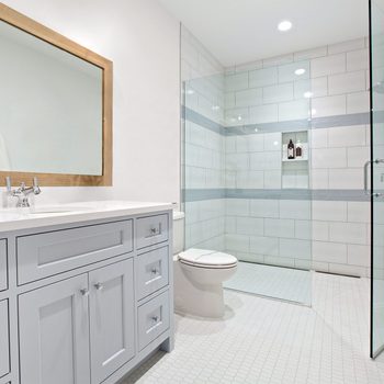 A modern bathroom featuring a white vanity with a wooden-framed mirror above it. There is a glass-enclosed walk-in shower with white and light gray tile. A white toilet is placed next to the shower. The floor is covered with small white tiles.