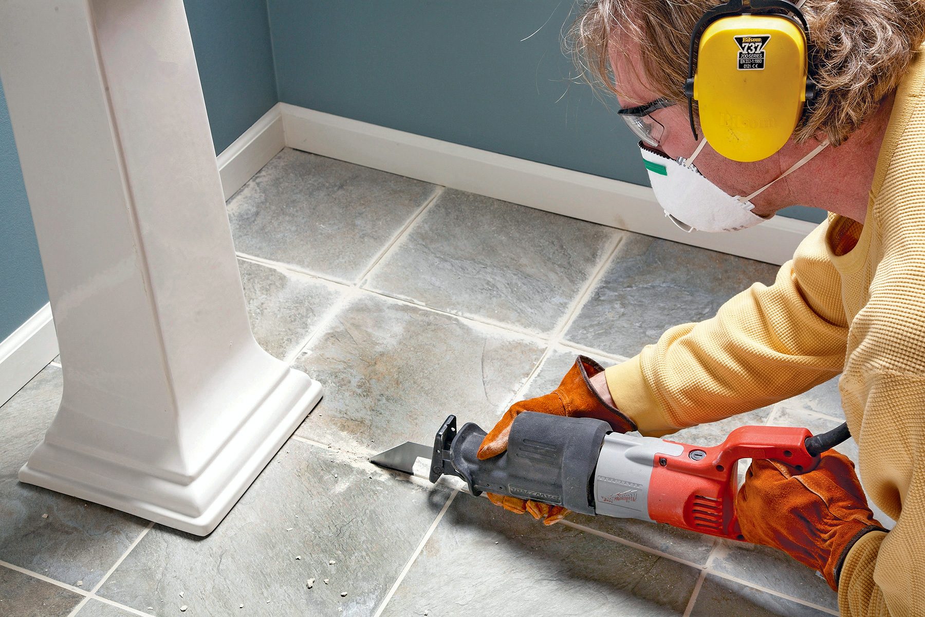 A person wearing safety goggles, ear protection, gloves, and a mask uses an electric saw to cut into a tiled floor near the base of a pedestal table. The person appears focused on the task in a corner of a room.